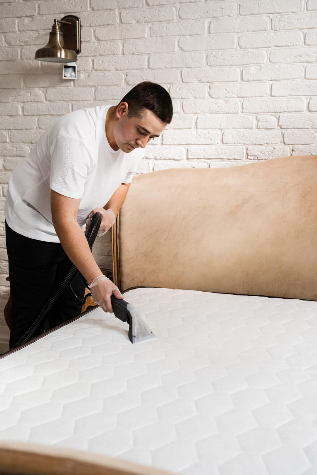 Homme nettoyant un matelas avec un aspirateur à main dans une chambre au mur de briques blanches.