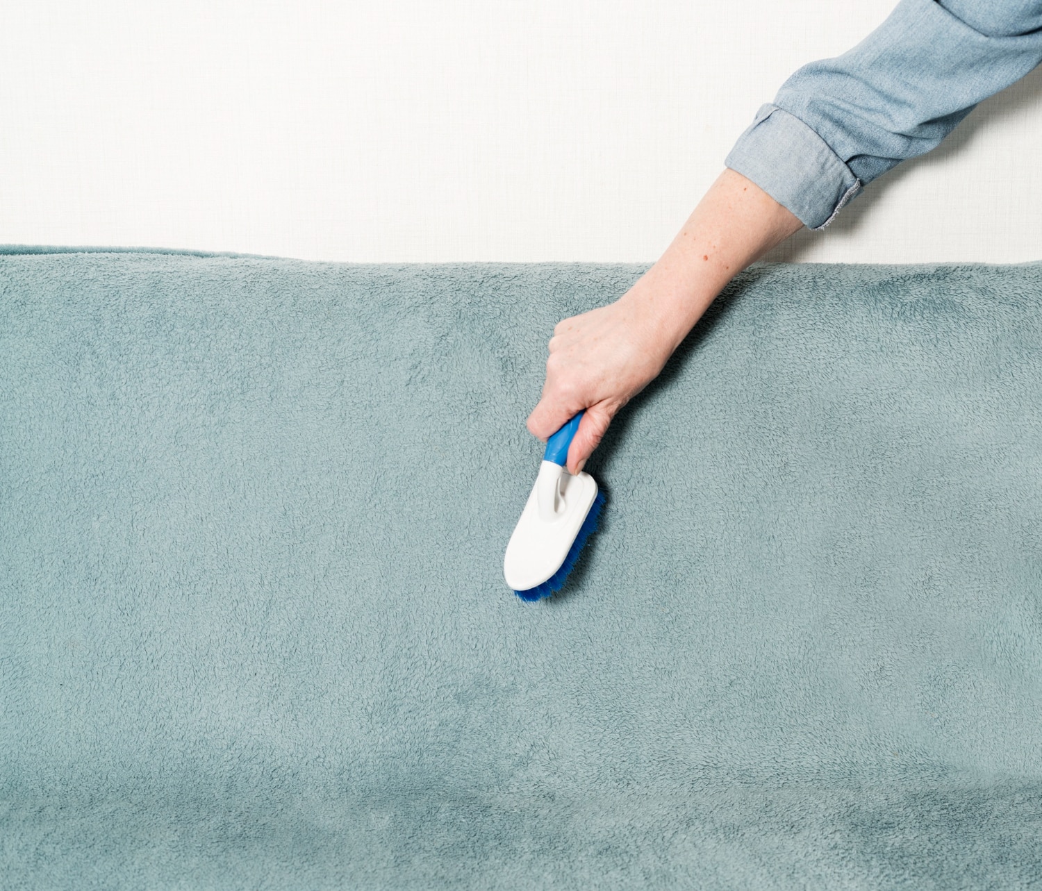 Main féminine brossant vigoureusement un tapis bleu ciel avec une brosse à main blanche à manche bleu.